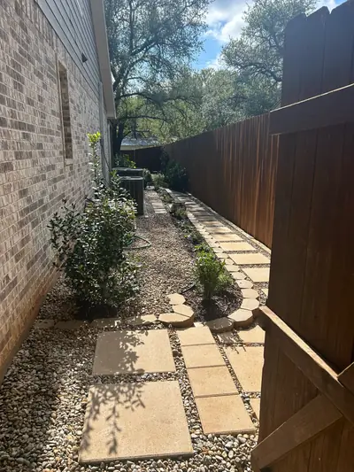 After: a curving stone path and gravel with shrubs beside the brick house and wooden fence.